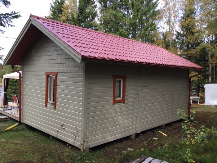 Nybyggd grå stuga med rött tak, nästan färdigställd utanför, omgiven av grönska.