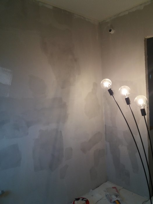 Vägg med ojämnt bredspacklat ytskikt och synliga lufthål under renovering, med arbetsbelysning och spackelredskap på golvet.