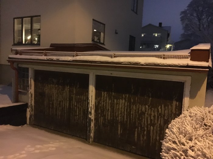 Snötäckt garage med slitna träportar framför ett bostadshus på kvällen.