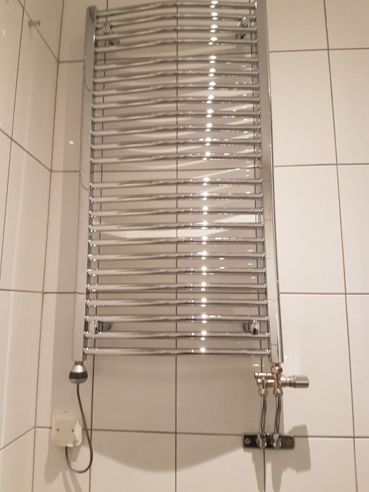 Handdukstork monterad på vit kakelvägg i badrum, med termostat och elektrisk anslutning synlig.