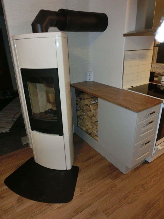 Vit kamin med svart rör syns bredvid köksskåp och ekgolv, ved staplad, svarta detaljer.