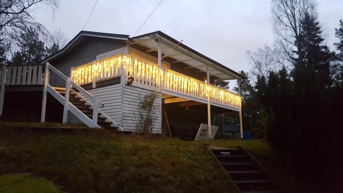 Ett litet hus från 1968 med upplyst veranda och trappa i skymningen.