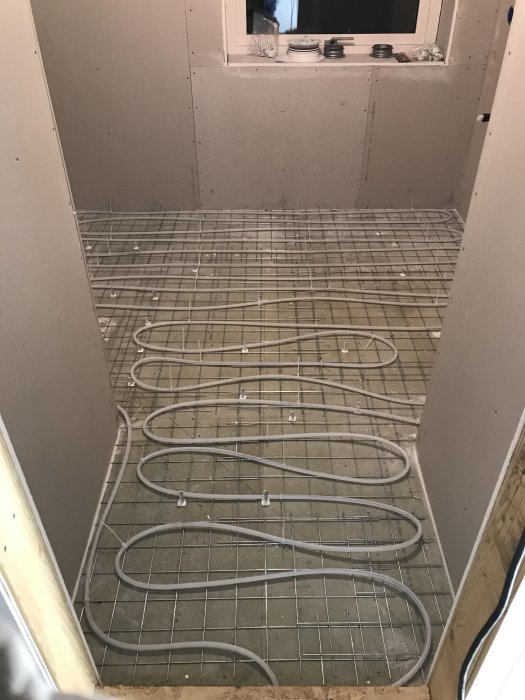 Golvvärmeslangar installerade på armeringsnät på ett rågolv med gipsväggar runtomkring under renovering.