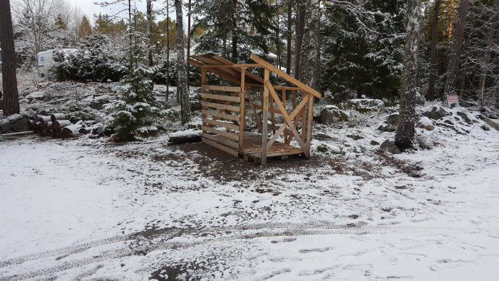 Tomt vedförråd i trä på snöig mark med träd och lätt snötäcke i bakgrunden.