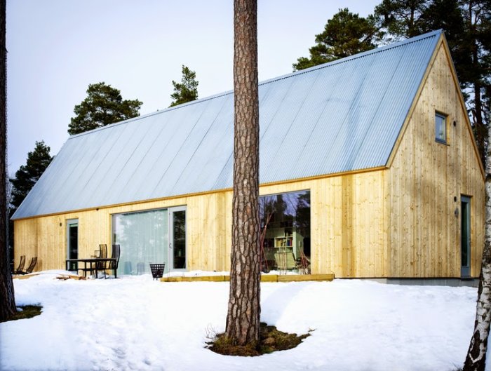 Modernt trähus i lada-stil med stora fönster, omgivet av snö och barrträd.