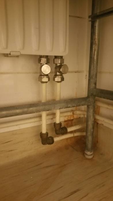Rör nära golv och väggen under en radiator med synliga genomföringar och avstängningsventiler.