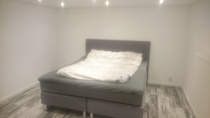 Renoverat sovrum med grå säng och ljust trägolv i en källare.