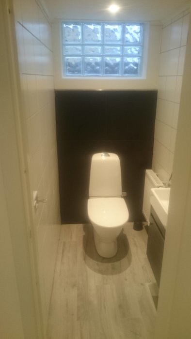 Renoverad källartoalett med vita kakelväggar, glasblocksfönster och mörkt baksäte.