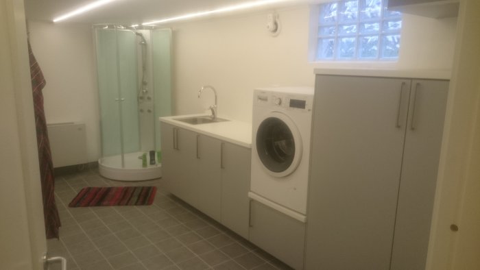 Renoverad källartvättstuga med duschhörna, tvättmaskin, handfat och förvaringsskåp.