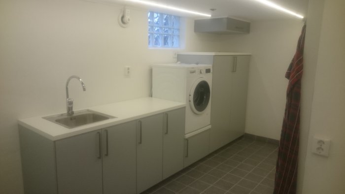 Nyrenoverad tvättstuga med vita skåp, tvättmaskin, handfat och litet fönster.