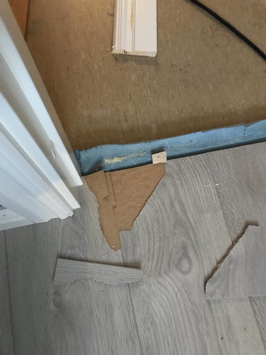 Skadat laminatgolv med uppstickande del nära dörrkarm och ersättningsbitar placerade på golvet.