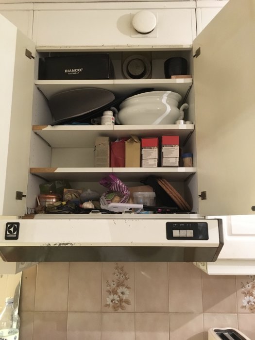 Köksfläkt med kolfilter ovanför kaklad vägg, frånluftsventil upptill, skåp med hushållsföremål.