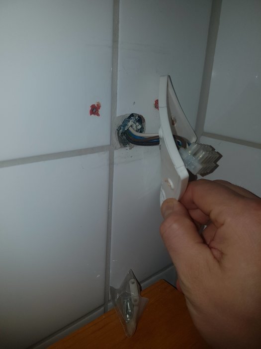 Hand håller ett doslock bredvid ett öppet kabelhål i vit kakelvägg i ett badrum, med synliga elektriska kablar.