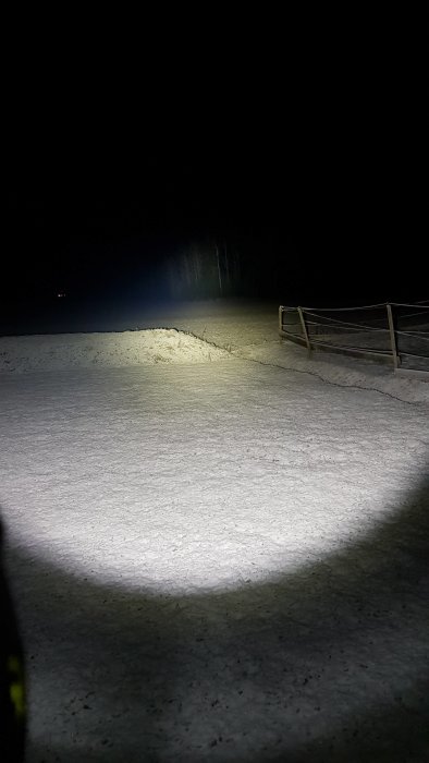 Kraftfull lampa lyser upp ett snötäckt fält och skog i mörkret, med ett smalt fokus på ljuset som når långt fram.