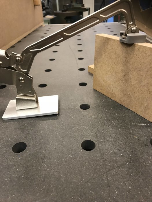 Metallklämma monterad på arbetsbänk som håller ett trästycke, visar klämman under spänning.