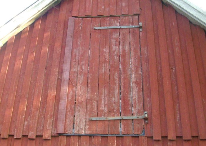 Vindslucka på rödfärgad träfasad med synliga gångjärn, behov av målningsarbete syns.