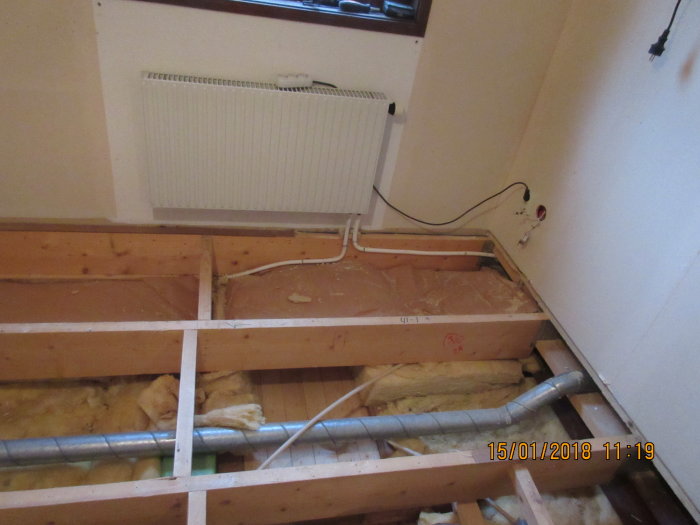 Rum under renovering med avlägsnat golv, synliga golvbjälkar, radiator, rörledningar och isolering.
