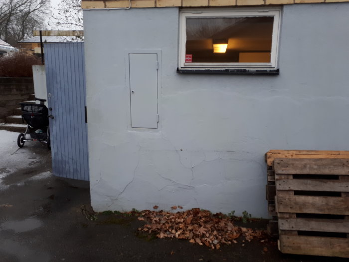 En vägg på ett garage med flagande färg och sprickbildning, ett fönster och en elektrisk panel.