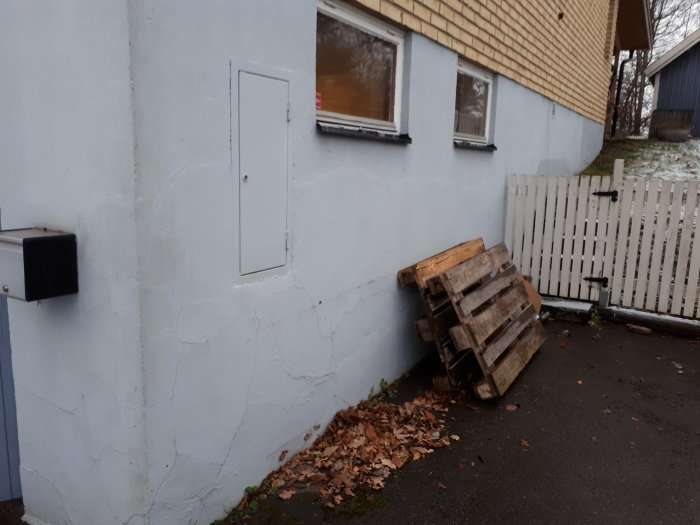 Yttervägg med flagande färg och spacklade borrhål, löv på marken, pall och vit staketdörr.