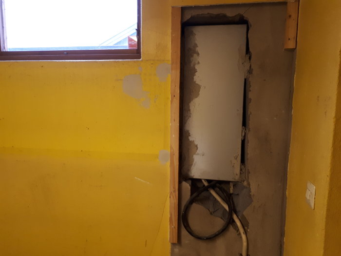 Elskåp med borttagen isolering i ett oisolerat och öppet utrymme på en gul vägg med synliga kablar.
