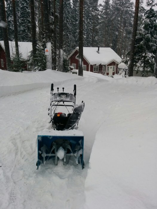 Snöslunga i drift framför röd stuga i ett snötäckt landskap.