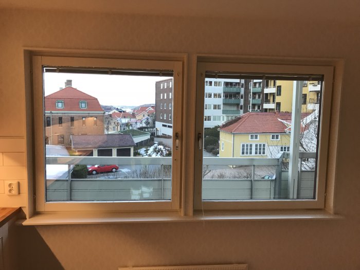 Utsikt från ett fönster som visar bostadshus, en röd bil och snöfläckar på marken.