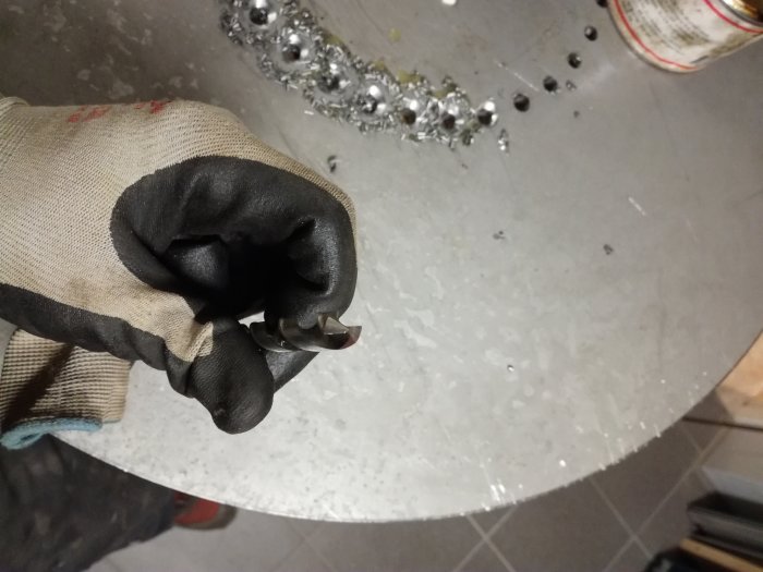 Hållen hand i skyddshandske håller ett använt 10 mm HSS borr framför en stålfot med borrade hål.