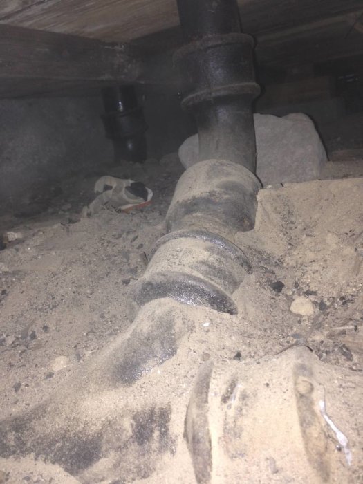 Äldre avloppsrör av gjutjärn i en dammig torpargrund, sannolikt under huset, med lite grus och stenar runtomkring.