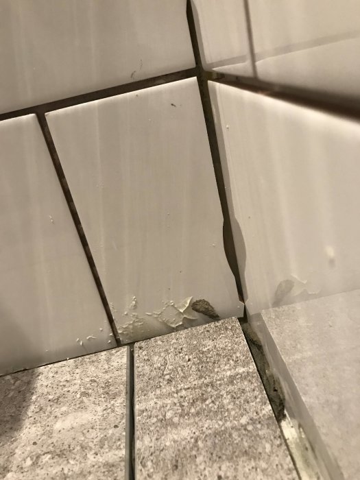 Hörn av ett badrum med ojämnt skurna kakelplattor, varierande fogbredd och glipa mot golvet.