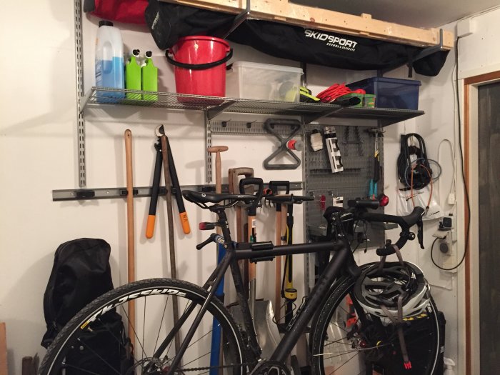 En nyinstallerad förvaringshylla med krokar och prylar i ett garage, inklusive cyklar, redskap och en vattenspridare.