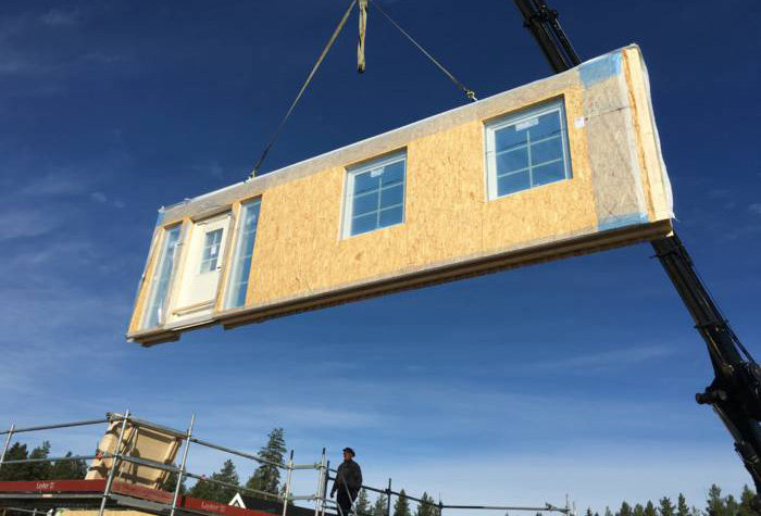 Prefabhussektion lyfts med kran vid husbygge, klarblå himmel i bakgrunden.