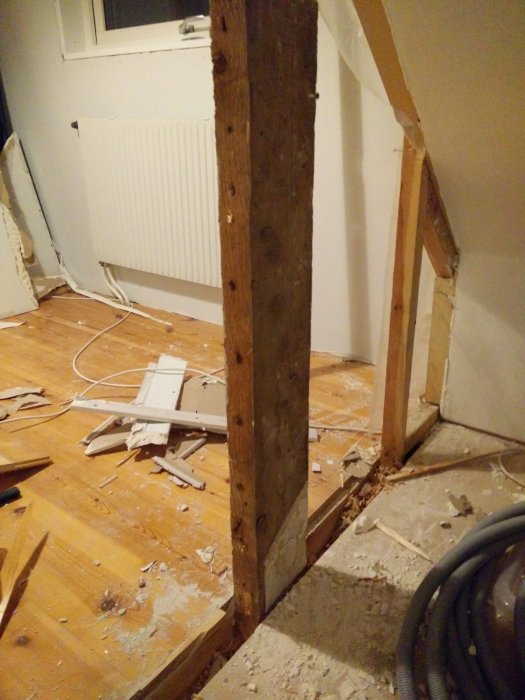 Övervåning under renovering med ett synligt stödben intill fönster och radiator, omgivet av byggavfall och nedrivna väggdelar.