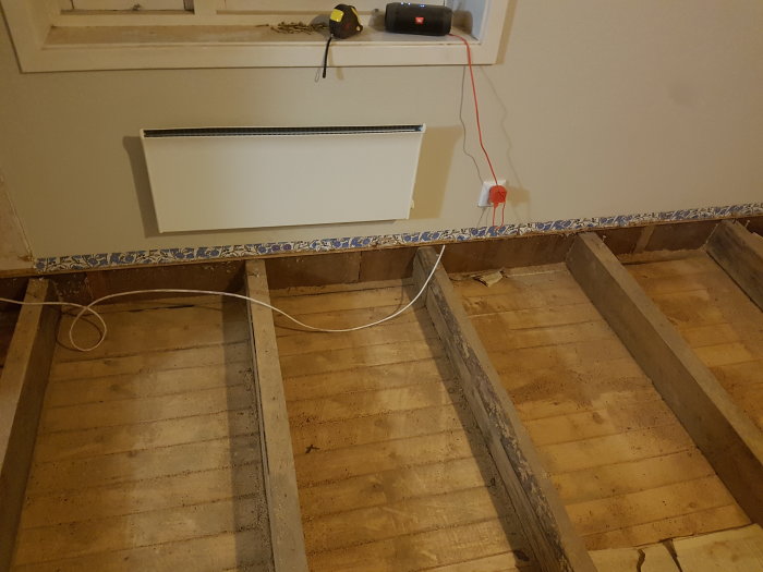 Delvis demonterat golv med exponerade träbalkar och en vägg med eluttag och en radiator, under en renovering.