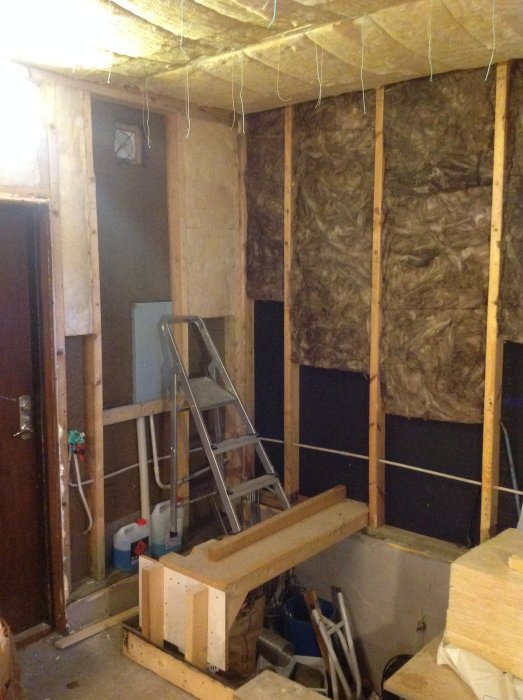 Garage under renovering med synlig isolering, trappstege och byggd plattform nära elskåp.
