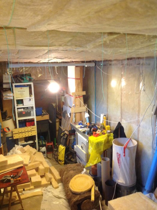 Oorganiserad verkstadsinteriör med trämaterial, verktyg och byggutrustning.