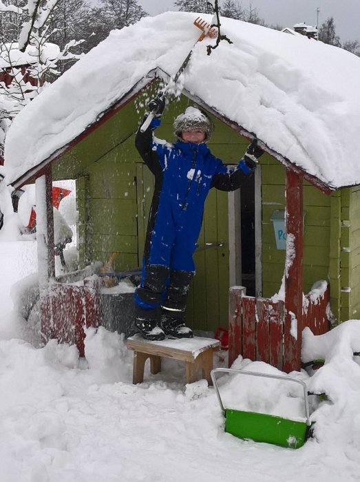 Person i blå overall som skottar snö från ett grönt lekhus tak.