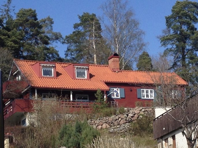 Rött hus med takkupor och Allmoge fönster under befintligt rött tak omgivet av träd.