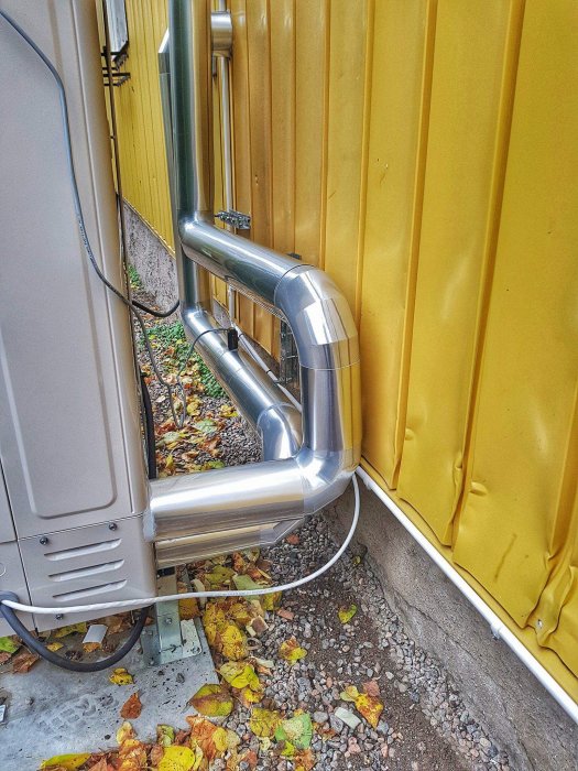 Värmepumps installation med välisolerade rör som löper längs en gul vägg och ner mot marken.