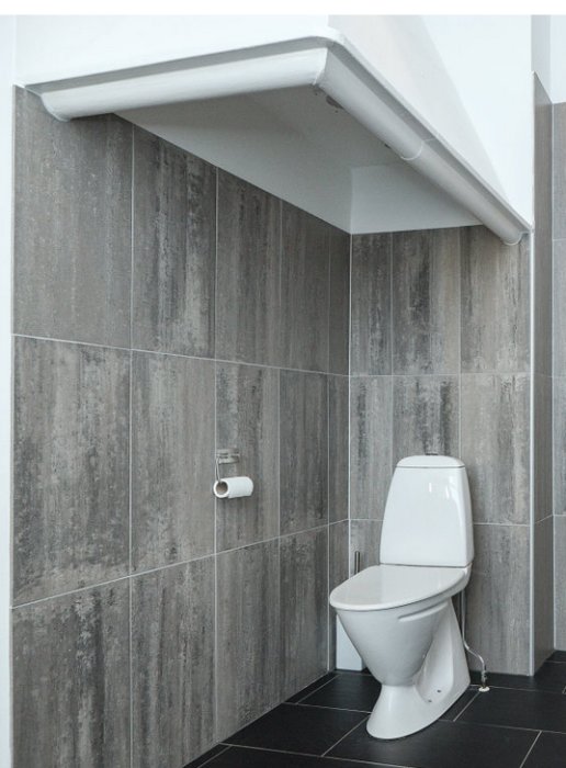 Toalett med ovanlig installation av vit ventilationskåpa överst på kaklad vägg.