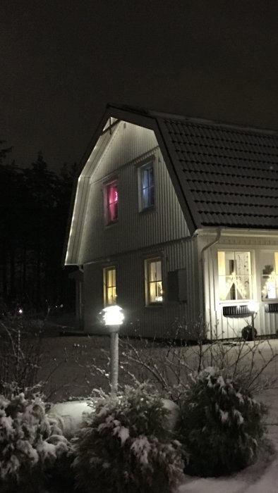 Ett hus på kvällen med snöklädda buskar och ett upplyst fönster i rosa nyans.