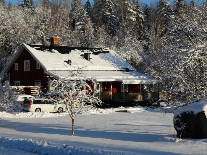 Röd stuga med snötäckt tak och omgivning i soligt vinterväder, med bil parkerad framför.
