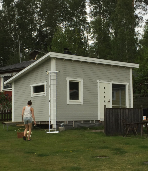 Person går med hink nära ett nyligen renoverat grått hus med vit dörr och fönsterfoder, stege lutad mot fasaden.