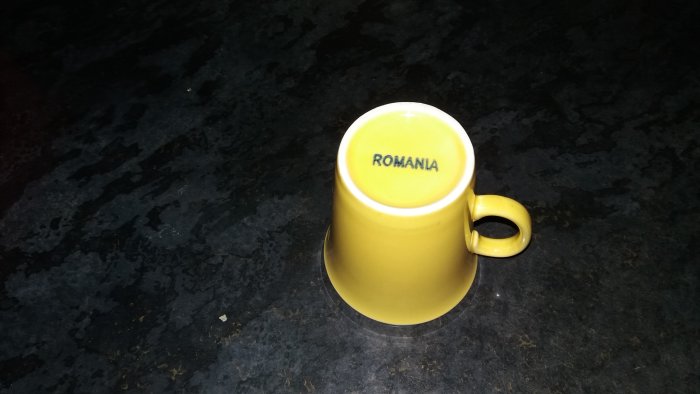Gul kaffemugg med texten "ROMANIA" på en mörk stenbänkskiva.