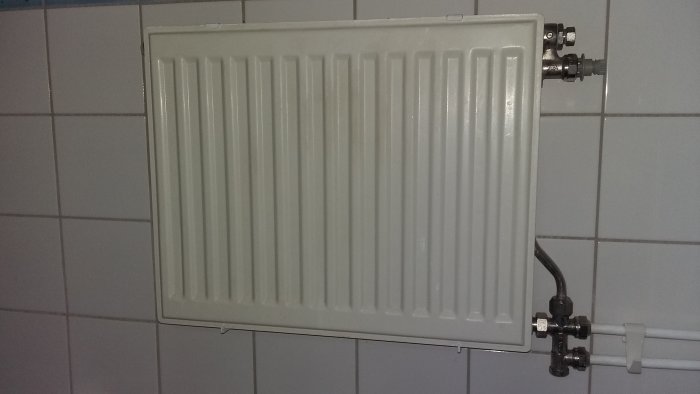 Vit radiator på kakelvägg, ser oanvänd ut med synliga ventiler och avstängningsventil.