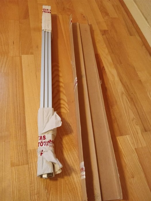 Aluminiumprofiler inslagna i hushållspapper och tejp, vid sidan av kartongemballage, på ett trägolv.