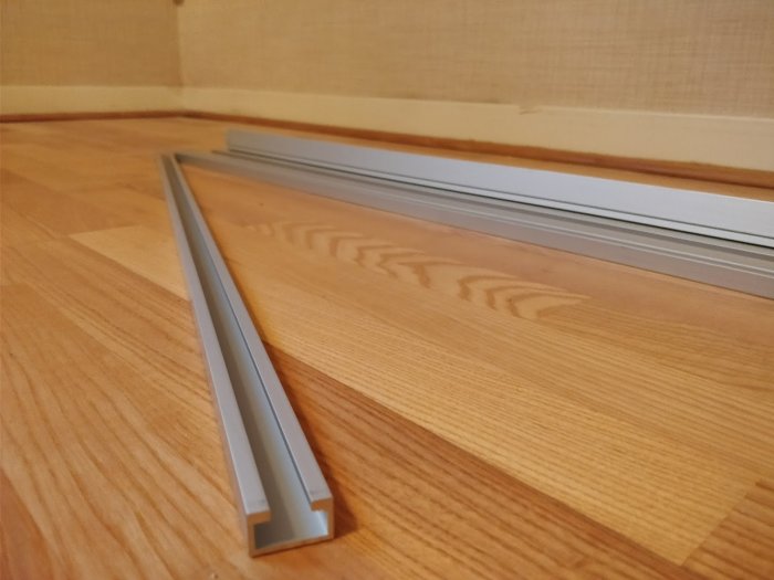 Aluminiumprofiler som ligger staplade på ett trägolv med en vit vägg i bakgrunden.