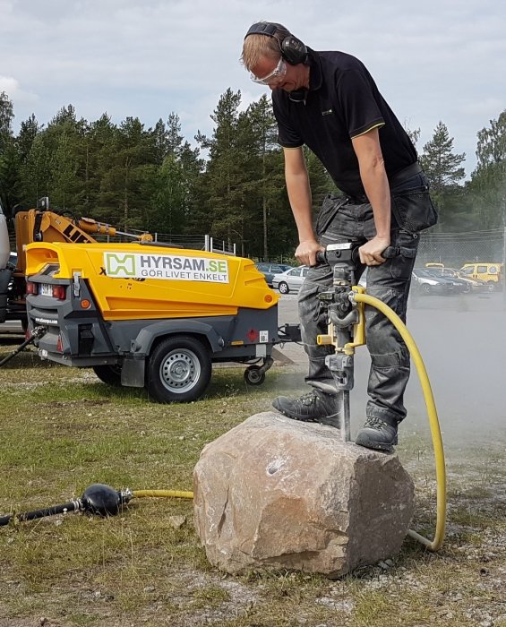 Person använder en bergborrmaskin medan kompressor på släpkärra är i bakgrunden.