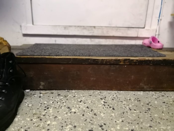 Höjdskillnad mellan garagegolv och tröskel med träram och stenplatta, terrazzo-mönstrat golv syns, rosa tofflor i bakgrunden.