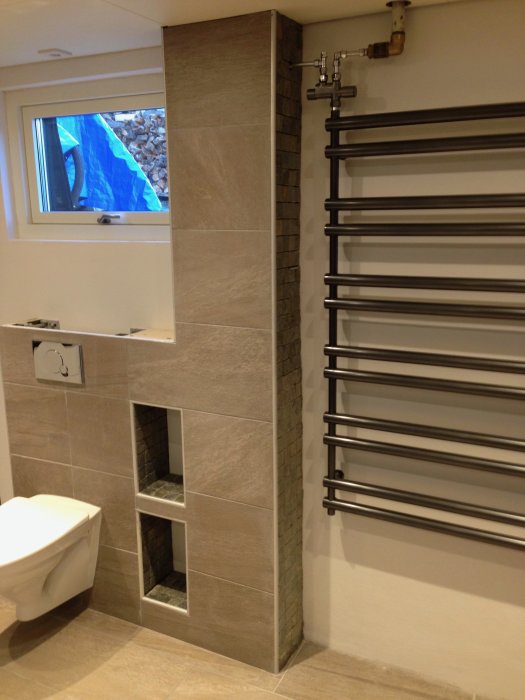 Ej färdigställd bastu med uppsatt panel, kaklade väggar och handdukstork intill ett väggmonterat toalett.