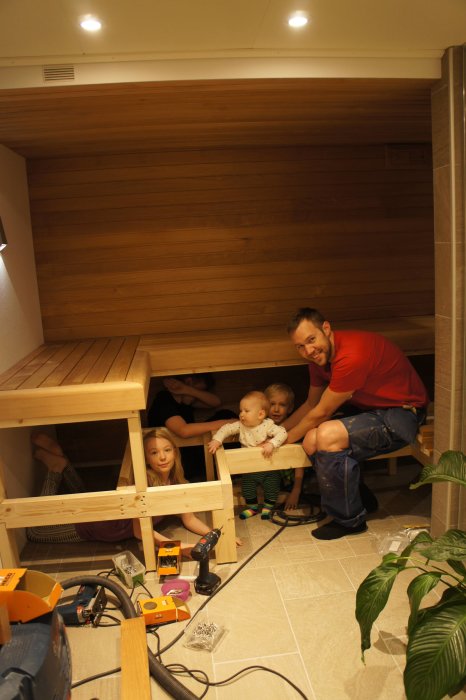 En person bygger bastu med barn, träväggar och bastulav, verktyg och material på golvet.